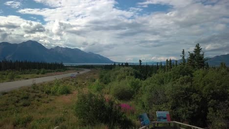 Panoramic-scene-by-official-Yukon-Kluane-lake-sign-of-car-driving-on-Alaska-highway-by-lake-towards-wild-rugged-Sheep-mountain-range-in-remote-countryside-landscape,-Canada,-overhead-rising-aerial