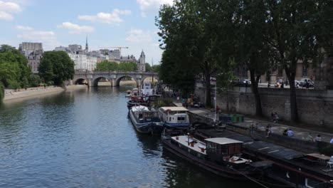 Paris-Seine-river-and-barges-boats-parked-by-the-quay-and-ile-de-la-cité-in-the-background-and-Pont-Neuf,-wide-slow-pan-shot