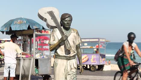 Statue-of-composer-Tom-Jobim-holding-a-guitar-over-his-shoulder-on-Ipanema-boulevard-with-street-vendors-and-ocean-in-the-background-and-people-in-face-mask-passing-by-walking-and-cycling