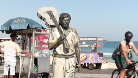 Estatua-Del-Compositor-Tom-Jobim-Sosteniendo-Una-Guitarra-Sobre-Su-Hombro-En-El-Bulevar-Ipanema-Con-Vendedores-Ambulantes-Y-El-Océano-En-El-Fondo-Y-Gente-Con-Mascarillas-Pasando-Caminando-Y-En-Bicicleta