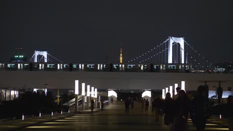 Escena-Nocturna-De-La-Pasarela-De-Odaiba-Con-Gente-Caminando,-Y-Un-Monorraíl-Pasando-Con-El-Puente-Del-Arco-Iris-Al-Fondo-En-La-Bahía-De-Tokio,-Tokio,-Japón