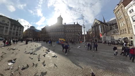 El-Palacio-Real-Con-Palomas-Y-Gente-En-La-Plaza-Dam-En-Amsterdam,-Países-Bajos-En-Una-Mañana-Soleada---Vista-De-Ojo-De-Pez,-Tiro-Panorámico