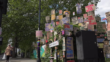 Un-Hombre-Con-Una-Máscara-Facial-En-Un-Scooter-De-Movilidad-Pasa-Un-Muro-Conmemorativo-De-Carteles-De-Cartón-De-Apoyo-Al-Personal-De-Atención-Médica-Durante-El-Brote-De-Coronavirus-En-El-Borde-De-Un-Parque-En-El-Extremo-Este-De-Londres