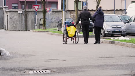 Blick-Hinter-Ein-älteres-Ehepaar,-Das-An-Einem-Wintermorgen-In-Arcore,-Norditalien,-Mit-Dem-Rollstuhl-Auf-Der-Straße-Spazieren-Geht