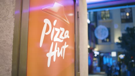 Cerrar-La-Toma-En-Cámara-Lenta-Del-Cartel-De-Pizza-Hut-Mientras-La-Gente-Borrosa-Camina-En-El-Fondo