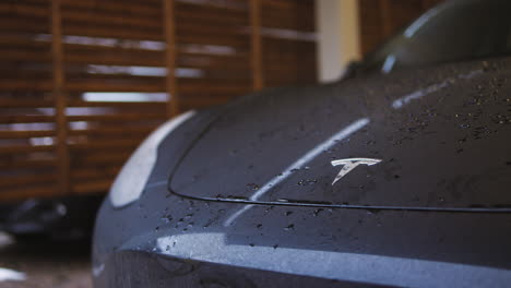 Langsame-Nahaufnahme-Des-Tesla-Logos-Auf-Der-Motorhaube-Eines-Nassen,-Dunklen-Autos-In-Der-Heimischen-Garage