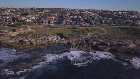 Flying-sideways-aerial-shot-of-ocean-waves-crashing-at-seashore-and-rockpool-near-waterfront-houses-and-beach