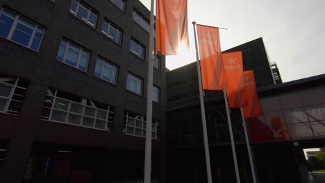 Dolly-shot-of-a-row-of-flags-with-the-MECC-name-and-logo-in-front-of-the-MECC-concert-venue-in-Maastricht,-the-Netherlands,-backlit-by-sunlight