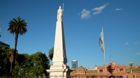 Bandera-Argentina-Ondeando-Contra-El-Cielo-Azul,-La-Pirámide-De-Mayo-Y-La-Casa-Rosada-En-La-Plaza-De-Mayo
