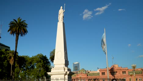 Argentinian-flag-waving-against-blue-sky,-May-Pyramid-and-Pink-house-in-May-Square