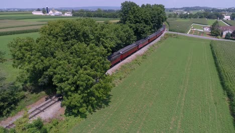 Una-Vista-Aérea-De-Una-Locomotora-Diésel-Tirando-De-Coches-De-Pasajeros-Antiguos-A-Través-De-La-Campiña-Amish