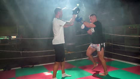 White-male-doing-kickboxing-practice-with-his-coach---punches-to-the-pads-while-running-on-the-spot,-slow-motion-video,-smokes-all-around,-camera-following-action
