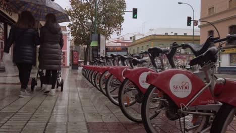 Frauen-Mit-Regenschirm-Schieben-Kinderwagen-Auf-Regnerischem-Bürgersteig-An-Leihfahrrädern-Vorbei