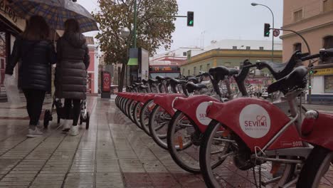 Women-with-umbrella-push-baby-stroller-past-rental-bikes-on-rainy-sidewalk