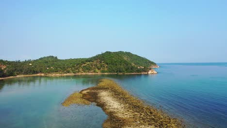 Pebbles-stripe-with-brown-algae-bordering-calm-lagoon-water-inside-bay-on-tropical-island-with-lush-vegetation-in-Thailand
