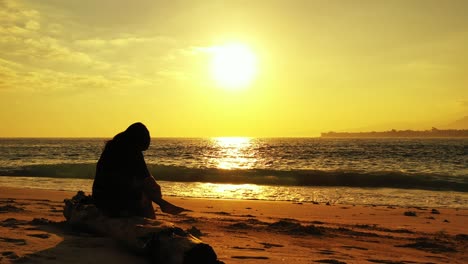 Silhouette-of-girl-sitting-on-dried-trunk,-watching-sun-setting-down-the-yellow-sky,-reflecting-on-sea-surface