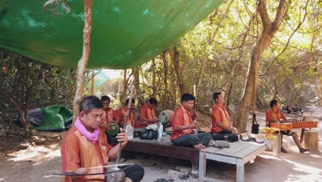 Weite-Außenaufnahme-Einer-Musikkapelle-Von-Herren,-Denen-Einige-Gliedmaßen-Fehlen,-Die-In-Einem-Waldgebiet-In-Der-Nähe-Von-Angkor-Wat-Sitzen-Und-Traditionelle-Instrumente-Spielen