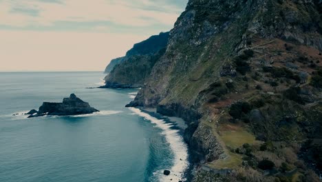 nobody,-aerial-birds-eye-view-of-a-contrast-seascape-with-isolated-rock,-volcanic-pebble-rock-beach-and-huge-mountain,-waves-and-white-foam-in-the-Atlantic-ocean-Daytime-at-Madeira-island-HD-landscape
