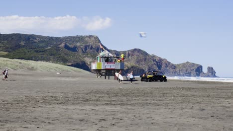 Surfers-walking-past-lifeguard-tower-at-Bethells-Beach,-West-Coast,-New-Zealand