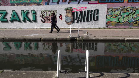 People-walk,-cycle-and-run-past-anti-Coronavirus-graffiti-art-of-a-character-wearing-a-surgical-face-mask-and-the-message-“stay-strong-and-don’t-panic”-on-a-wall-in-East-London