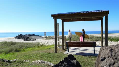 Surfer-Holding-A-Surfboard-On-The-Beach---Beachfront-Gazebo-With-Wooden-Bench---Gold-Coast,-Queensland,-Australia