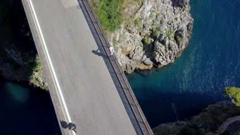 Puente-De-Arco-Fiordo-Di-Furore-Con-Un-Hombre-Con-Sombrero-Admirando-La-Vista-Mientras-Pasan-Los-Autos,-Toma-Aérea-De-La-órbita-Superior-Del-Dron