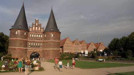Toma-Panorámica-Derecha-Del-Famoso-Edificio-De-La-Puerta-De-Holsten-Con-Turistas-Visitantes-Durante-La-Puesta-De-Sol-Por-La-Noche-En-Lübeck,-Alemania