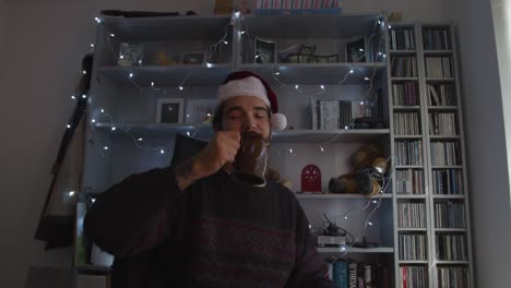 Hombre-Hablando-A-La-Cámara-En-Una-Videollamada,-Brindando-Con-Una-Taza-Mientras-Usa-Un-Sombrero-De-Navidad