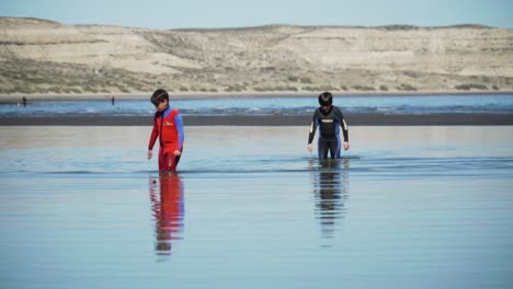 Dos-Niños-Con-Trajes-De-Neopreno-Caminando-En-Las-Aguas-Poco-Profundas-Del-Mar-Patagónico,-Fondo-De-Montaña---Tiro-Ancho-Uhd