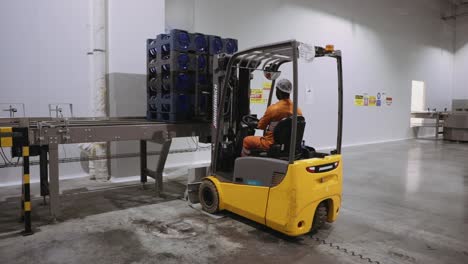 Masked-worker-moving-water-gallons-crates-using-forklift-in-warehouse