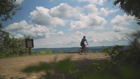 Static-shot-slow-motion-of-young-man-riding-mountain-bike-on-off-road-trail-overlooking-bright-blue-sky-with-white-clouds-in-a-forest-in-Pennsylvania-in-the-summer