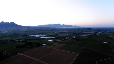 Aerial-drone,-dusk-sunrise-over-farm-land-close-up-of-brown-vineyards,,-ponds,-dams-and-vineyards,-light-blue,-pink-and-purple-sky-with-mountains