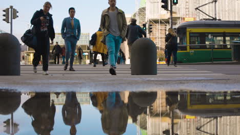 Vista-En-Cámara-Lenta-De-Una-Multitud-De-Personas-Cruzando-La-Calle-En-Finlandia-Con-Agua-Atrapando-Su-Reflejo