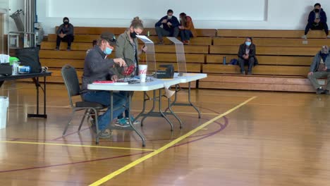 Man-wearing-face-mask-sits-at-a-desk-in-a-school-gymnasium-spinning-a-raffle-drum-and-pulls-a-lottery-number-to-hike-The-Wave