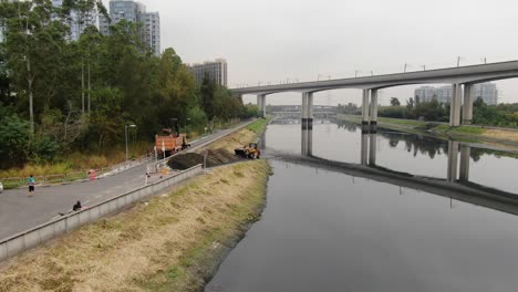 Gelber-Traktor,-Der-Schlamm-Aus-Einem-Kanal-Unter-Einer-Brücke-Schöpft-Und-Reinigt,-Luftbild