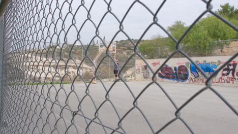 Vista-Desde-Detrás-De-Una-Valla-Metálica-De-Un-Hombre-Jugando-Baloncesto-En-Una-Cancha-Al-Aire-Libre