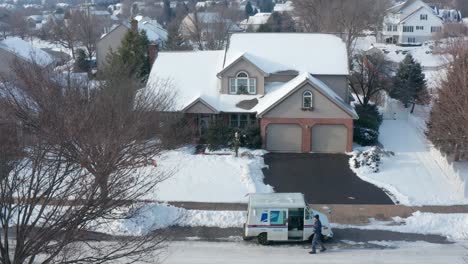 Usps-Mail-Man-Deja-Caer-Un-Paquete-De-Regalo-De-Navidad-En-Una-Casa-Cubierta-De-Nieve-Durante-La-Temporada-De-Vacaciones-De-Invierno-De-Navidad,-Vista-Aérea-De-Drones