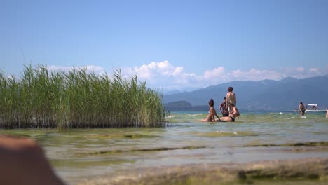 Wide-shot-of-people-sunbathing-on-the-rocks-in-shallow-lake-water-on-Jamaica-Beach,-Sirmione,-Lago-Garda-Lake-Garda,-Italy