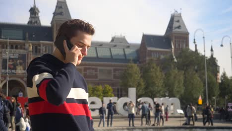 Handsome-young-man-talking-on-cellphone-and-enjoying-a-sunny-day-in-front-of-the-I-Amsterdam-sign-in-Amsterdam