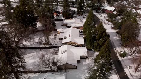 Aerial-Shot-of-Small-Rural-Town,-Rooftops-Covered-in-Snow-Surrounded-by-Pine-Trees-in-Winter