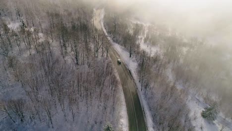 Aerial-view-of-Car-driving-on-winter-country-road-in-snowy-forest-covered-with-golden-fog-lit-up-by-sun-revealing-the-mountain-in-the-distance