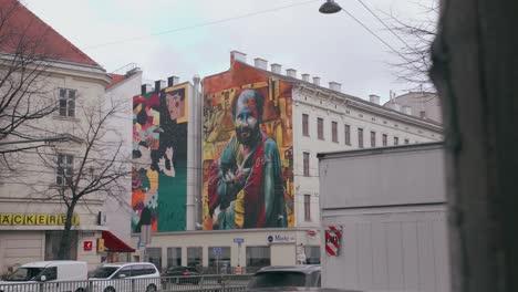 Colorful-street-art-on-a-whole-side-of-a-building,-in-the-middle-of-Vienna,-Austria,-with-vehicles-and-cars-passing-by,-near-other-buildings,-on-a-cloudy-day