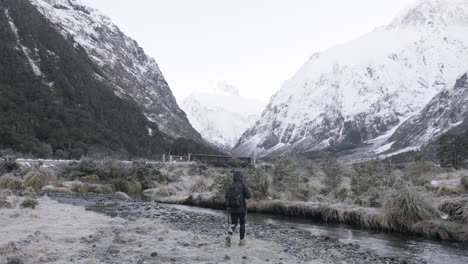 Slow-motion-shot-of-girl-in-hiking-gear-photographing-snow-capped-mountains-next-to-running-river-in-winter