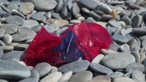 Bolsa-De-Plástico-Roja-Que-Ensucia-Una-Playa-De-Guijarros.-Zoom-Lento.