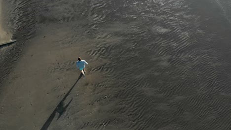 Aerial-top-down-tracking-shot-of-young-caucasian-man-running-on-a-beach-in-Auckland,-New-Zealand
