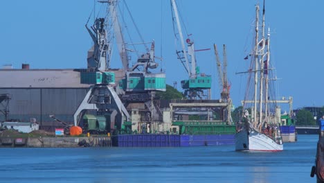 Sailing-vessel-Zawisza-Czarny-leaves-Port-Of-Liepaja-in-hot-sunny-summer-day,-port-cranes-in-background,-medium-shot-from-a-distance