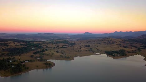 Fast-rising-aerial-view-of-lake-Moogera,-brown-hills-and-mountains-at-sunset---Queensland,-Australia