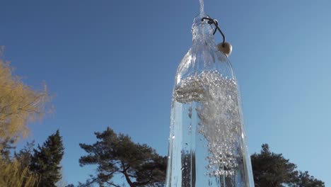 Pouring-water-in-glass-bottle-with-blue-sky-and-tree-in-background,-close-up,-low-angle