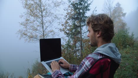 Mittlere-Aufnahme,-Seitenansicht-Eines-Mannes,-Der-Einen-Laptop-Benutzt,-Neblige-Sicht,-Bäume-Und-Gras-Im-Hintergrund