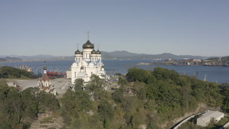 Langsamer-Abstieg-Luftaufnahme-Einer-Orthodoxen-Kirche-Mit-Blauem-Dach-Und-Goldenen-Kuppeln-Mit-Hafen-Und-Bucht-Im-Hintergrund-An-Einem-Hellen,-Sonnigen-Tag